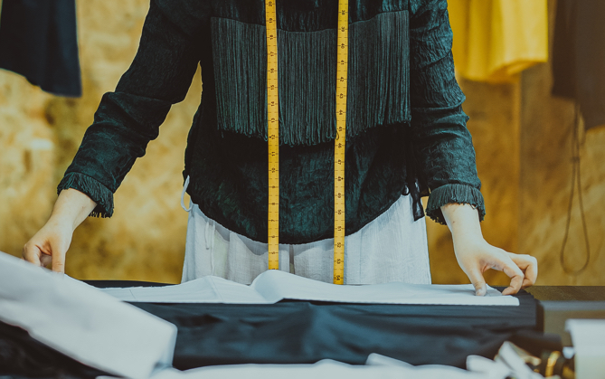  A person with measuring tape hanging from their neck, bent over a table laid with sheets of fabric.