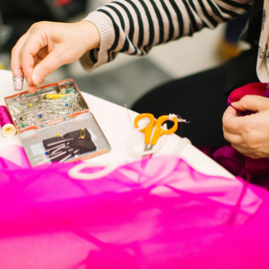 A person reaching for a box of sewing pins sitting on a table with pink fabric and scissors.